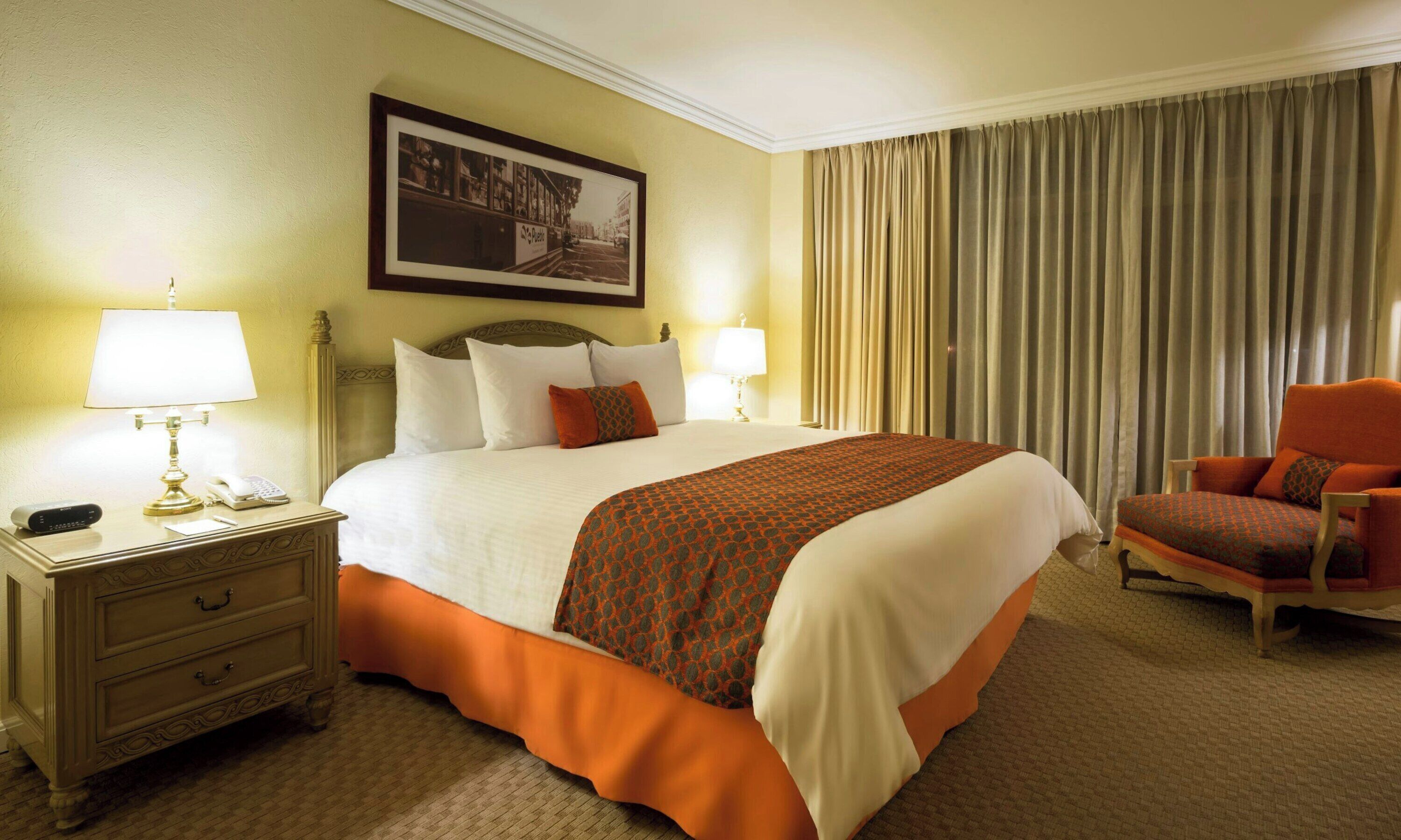 A hotel room with a large bed, two bedside lamps, a nightstand with a phone, a chair, and a framed picture on the wall.