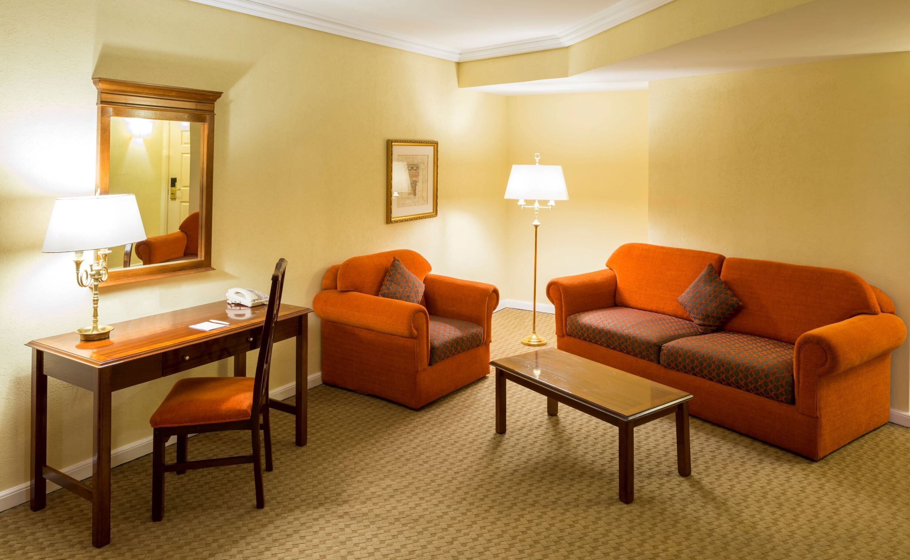 A hotel room with a wooden desk, two orange sofas, a small table, a lamp, a chair, and a mirror.