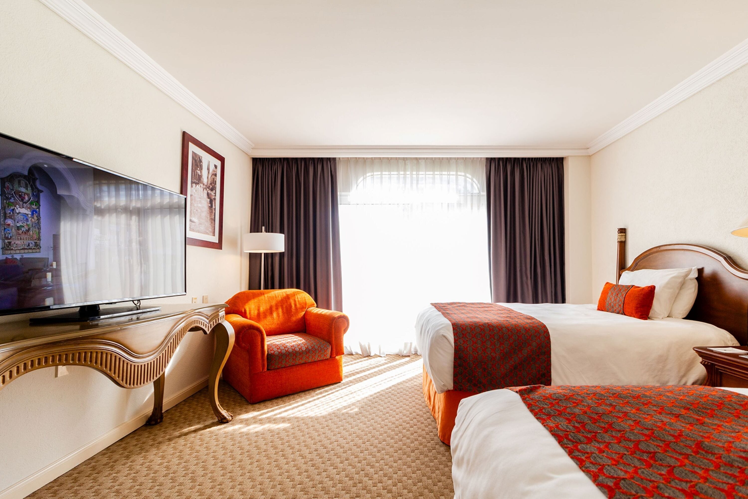 A hotel room with a television, a sofa, two beds, and a large window with curtains.