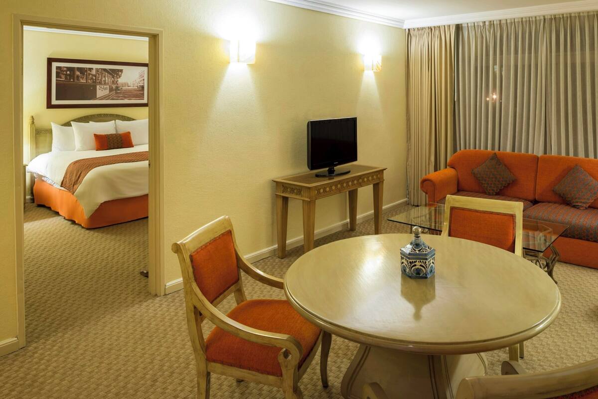 Une chambre d’hôtel avec un lit, un téléviseur posé sur un meuble en bois, un canapé orange, une table à manger avec des chaises et un miroir qui reflète la pièce.