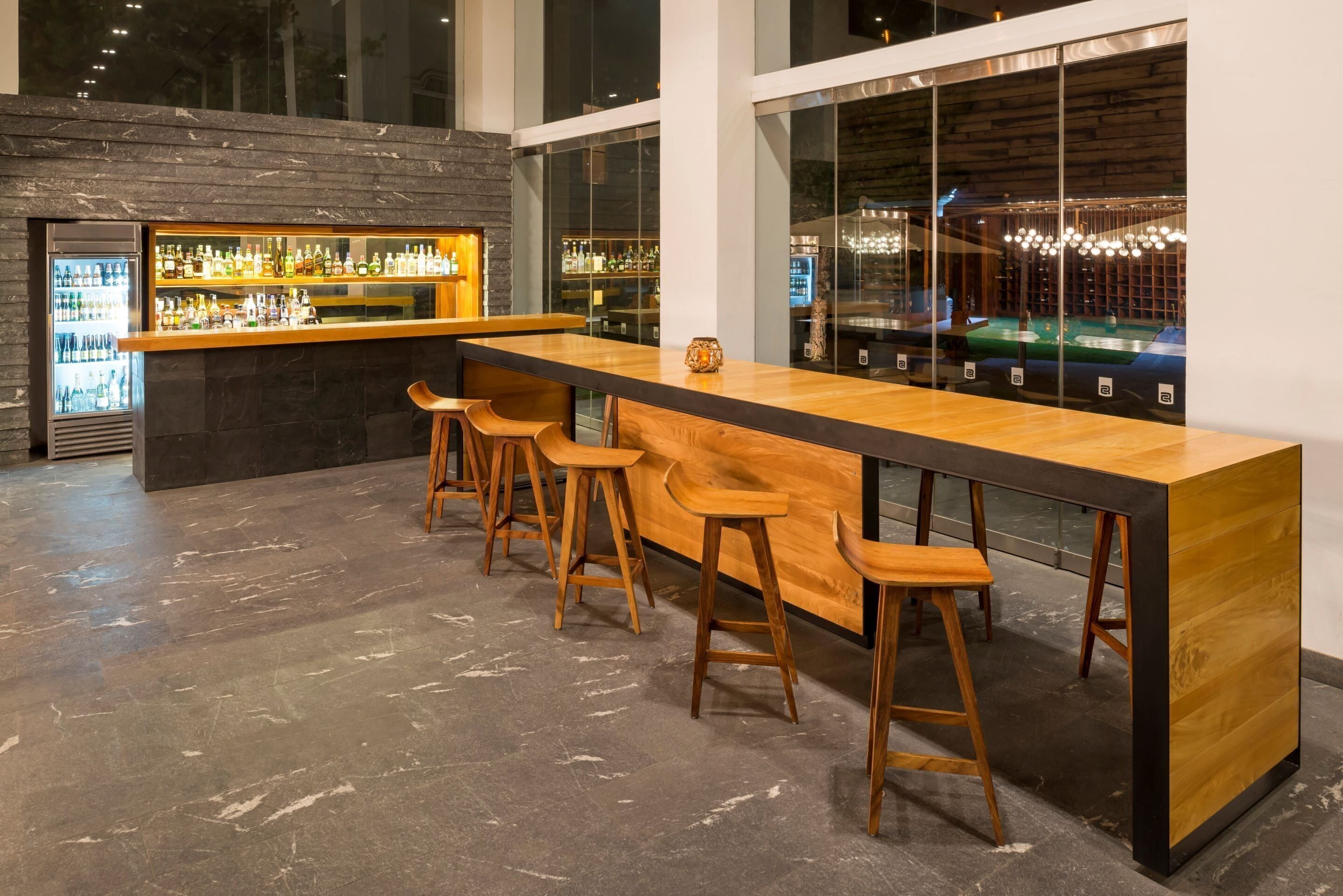 A bar area with a long counter, wooden stools, and a refrigerator stocked with beverages.