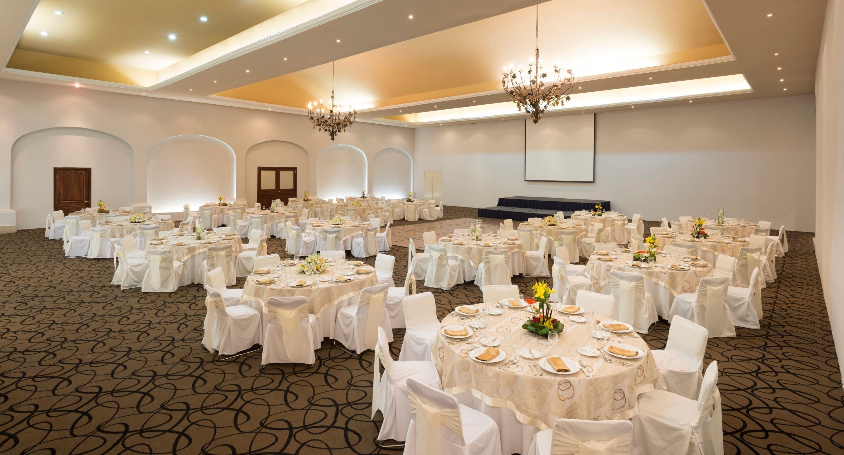 A large banquet hall with round tables, white chairs, and chandeliers.