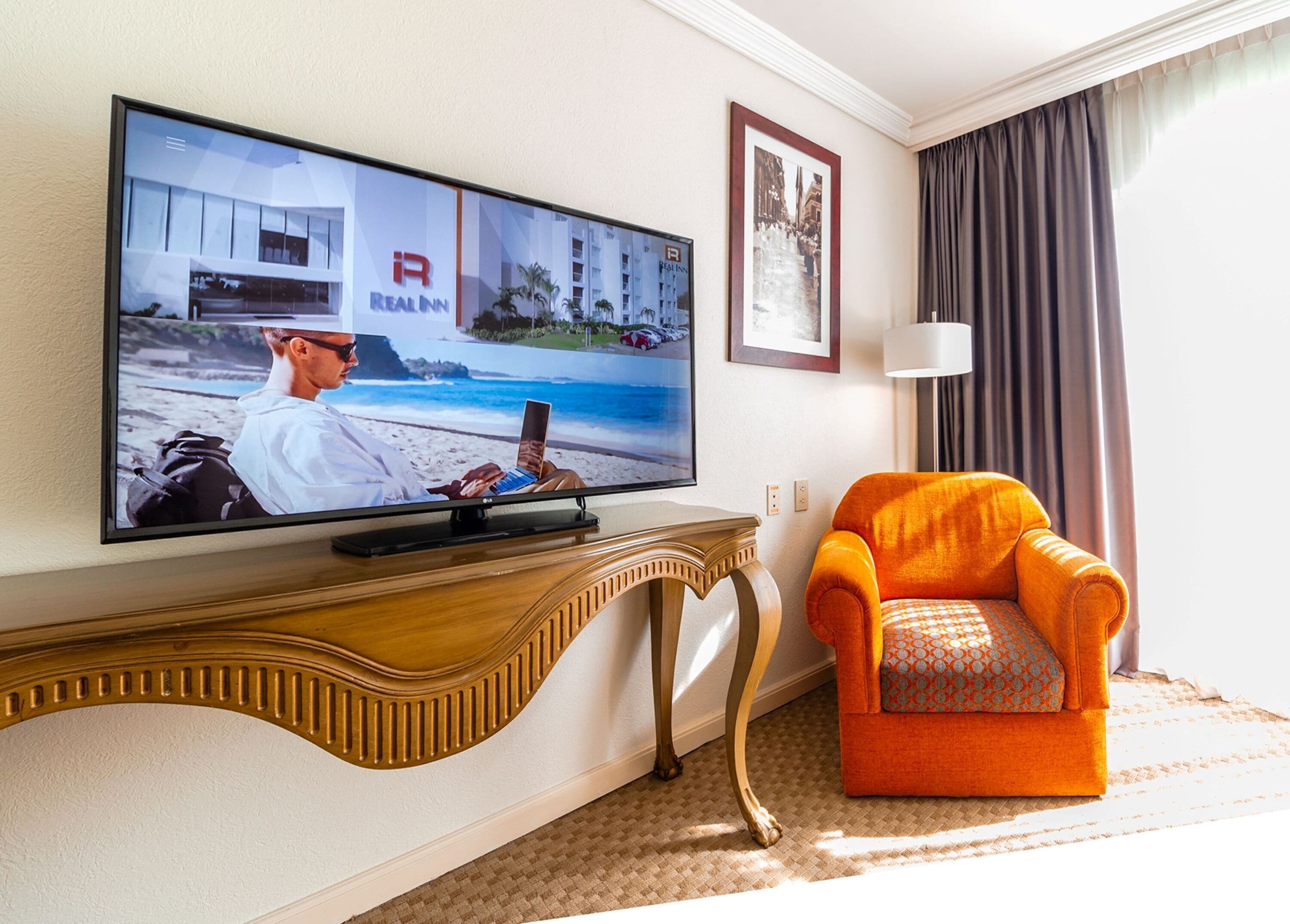 A hotel room with a large flat-screen TV mounted on a wooden console, an orange armchair, a framed picture on the wall, and a window with curtains.