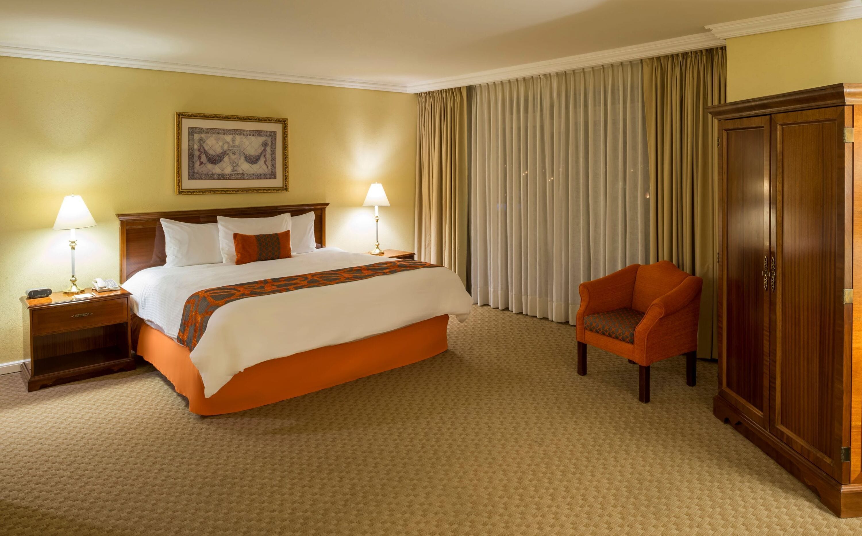 A hotel room with a large bed, two bedside lamps, a chair, a wardrobe, and a painting on the wall.