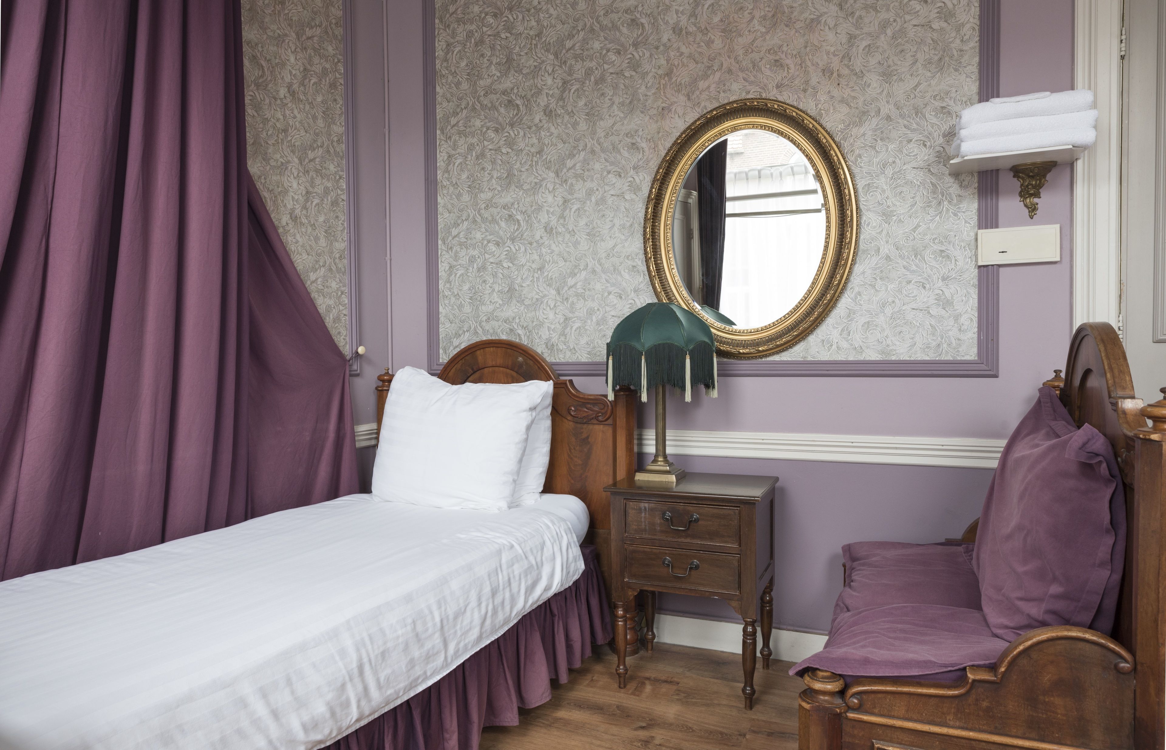 A bedroom with a bed, a nightstand, a chair, a mirror, and a wall with patterned wallpaper.