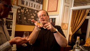 A person holding a glass of whiskey in a bar setting with a whiskey-themed wall and shelves of bottles. - Hotel Fidder - Patrick's Whiskybar (Zwolle)