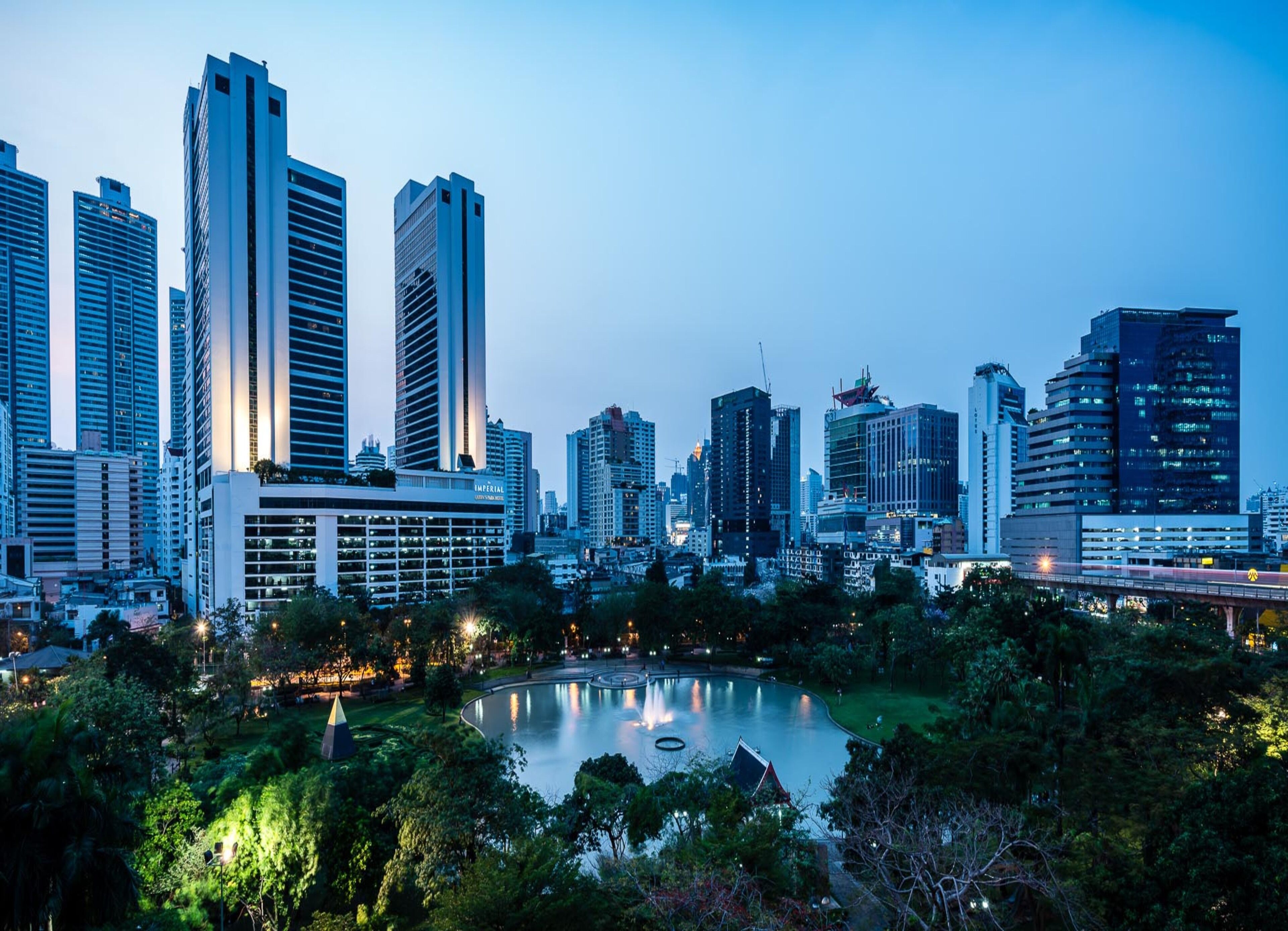 A cityscape with modern high-rise buildings, a central park with a pond, and a bridge in the foreground.