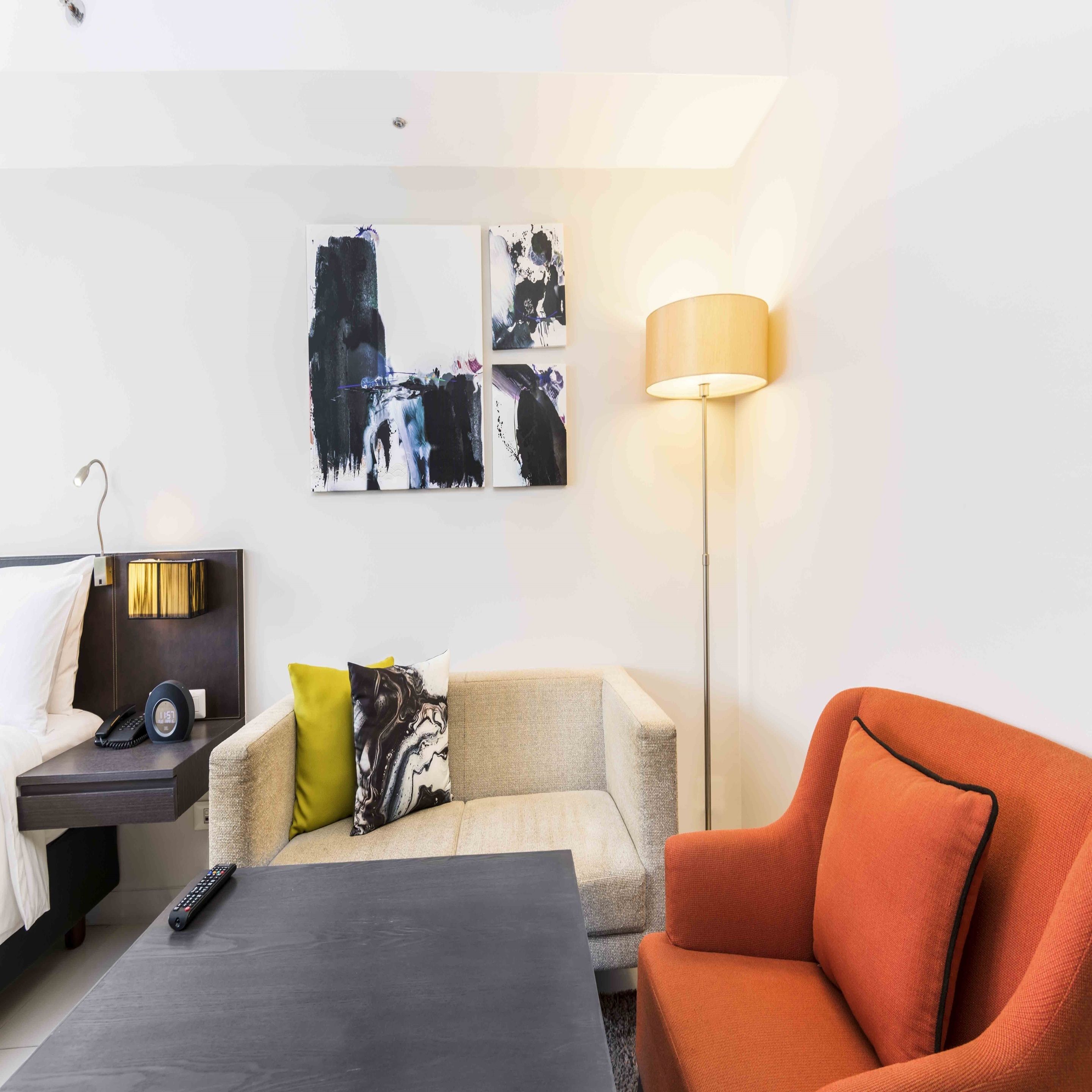 A hotel room with a bed, a desk with a lamp, a sofa, an orange armchair, and abstract art on the wall.