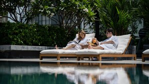 Zwei Personen entspannen auf Liegestühlen am Pool mit Blick auf üppiges Grün und ein Gebäude im Hintergrund.