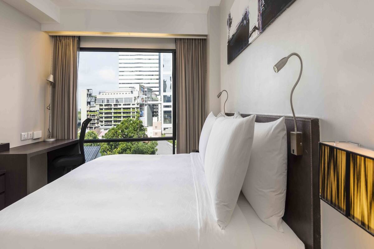 Habitación de hotel con una cama grande, un escritorio, una silla, una lámpara y vistas a edificios a través de la ventana.