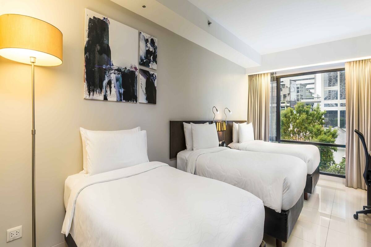 Habitación de hotel con dos camas, una lámpara de pie, arte abstracto en la pared y vistas de la ciudad a través de amplias ventanas.