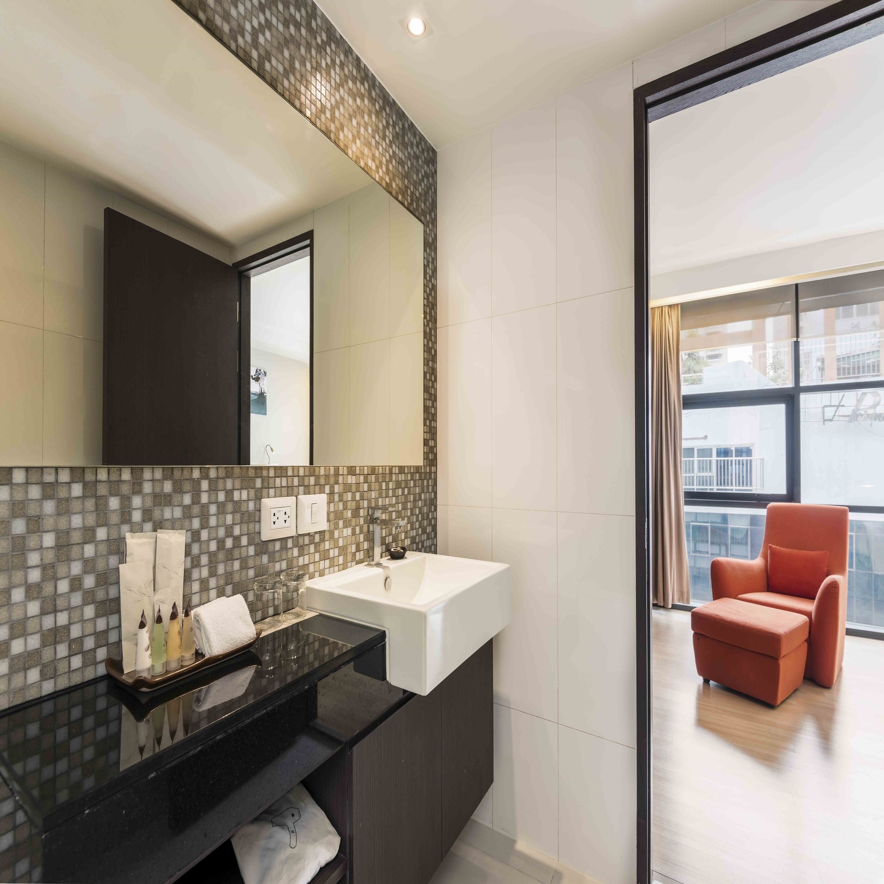 A modern bathroom with a large mirror, a white sink, and a black countertop. There is a window with a view of a balcony and a red chair.