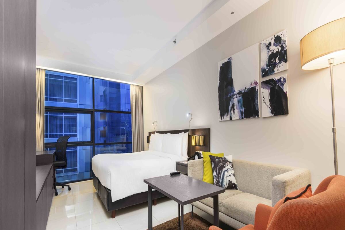 Habitación de hotel moderna con una cama grande, un escritorio, un sofá y una mesa de centro. Cuenta con un ventanal con cortinas, una obra de arte en la pared y una lámpara.