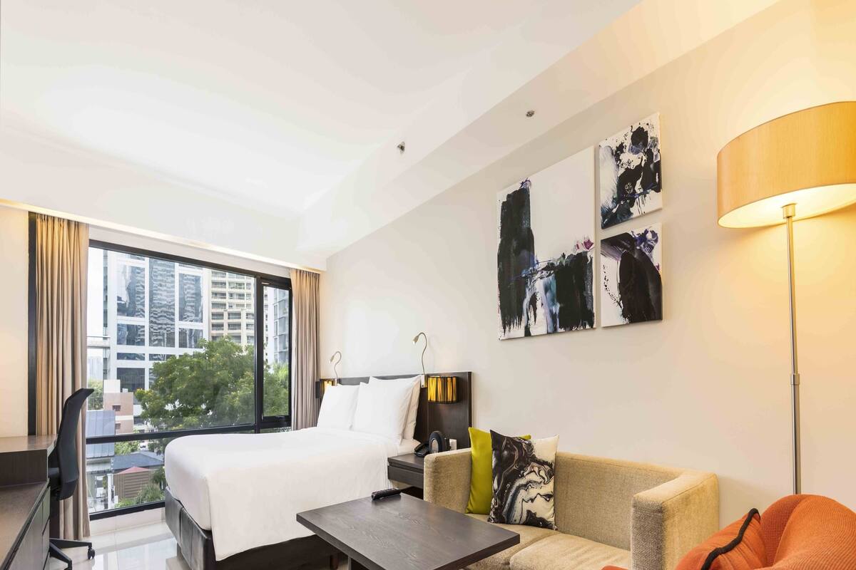 Una habitación de hotel moderna con una cama grande, un sofá, una mesa de centro y un escritorio. Se ve una vista de edificios a través de la ventana.