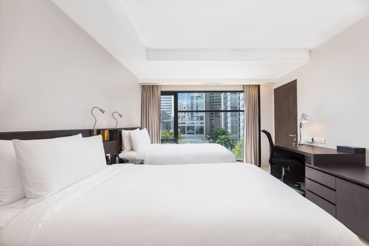 Habitación de hotel con dos camas, un escritorio y vistas a edificios a través de amplias ventanas.