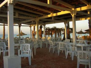 An outdoor seating area with white plastic chairs and tables, a tiled floor, and a view of the sea. - Regency Hammamet (Hammamet)