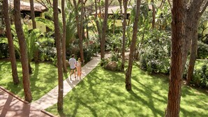 A pathway through a lush garden with trees and greenery, leading to a building with a tiled roof. - Forte Village Resort – Hotel Pineta (Pula)