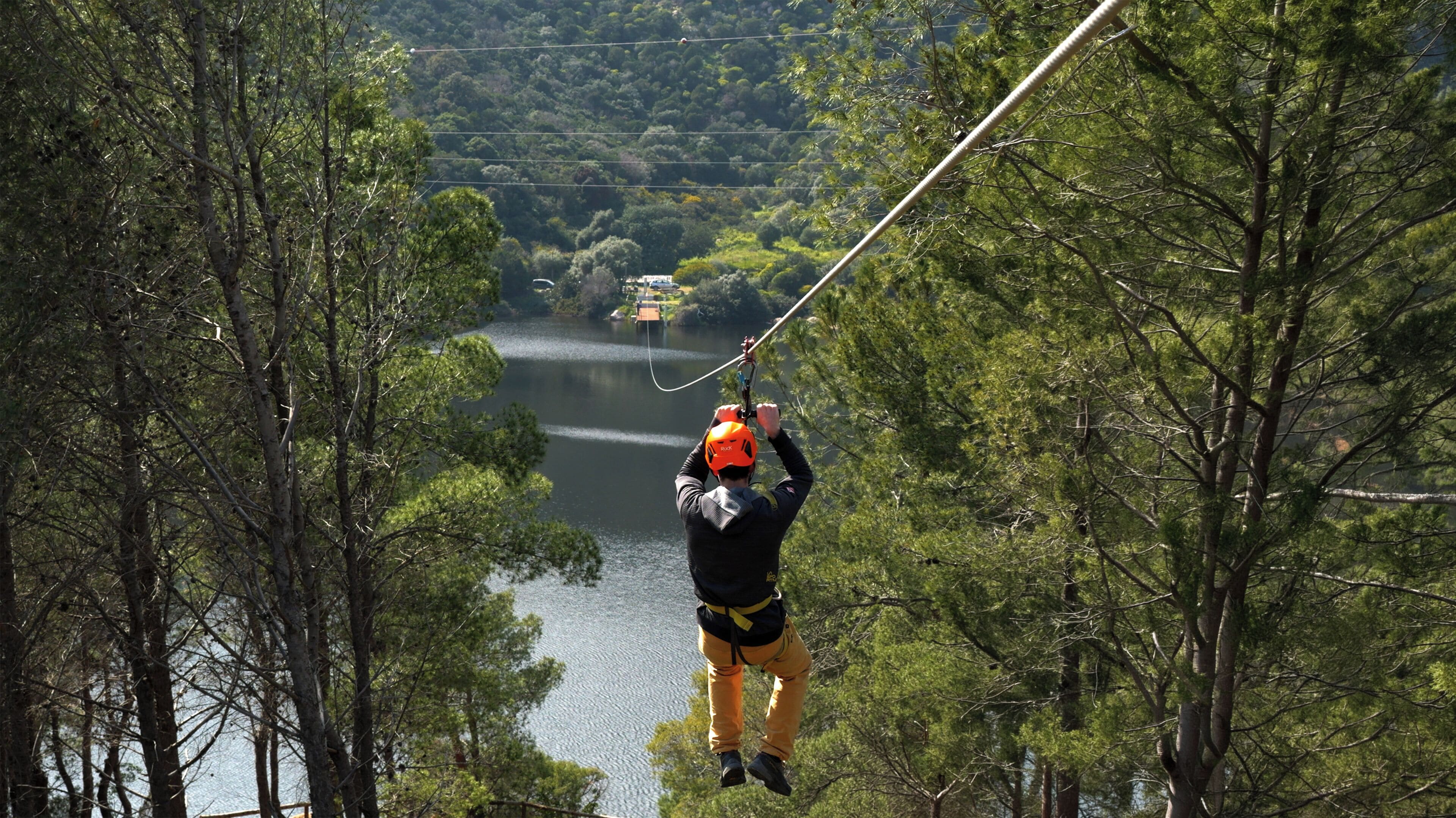 Une personne pratiquant le saut à l’élastique avec un harnais et un casque, entourée d’arbres et d’une étendue d’eau.