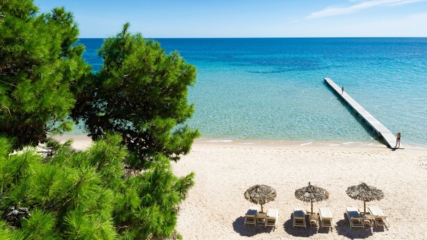 A beach with clear blue water, a pier extending into the sea, and several lounge chairs with umbrellas. - Forte Village Resort – Hotel Pineta (Pula)