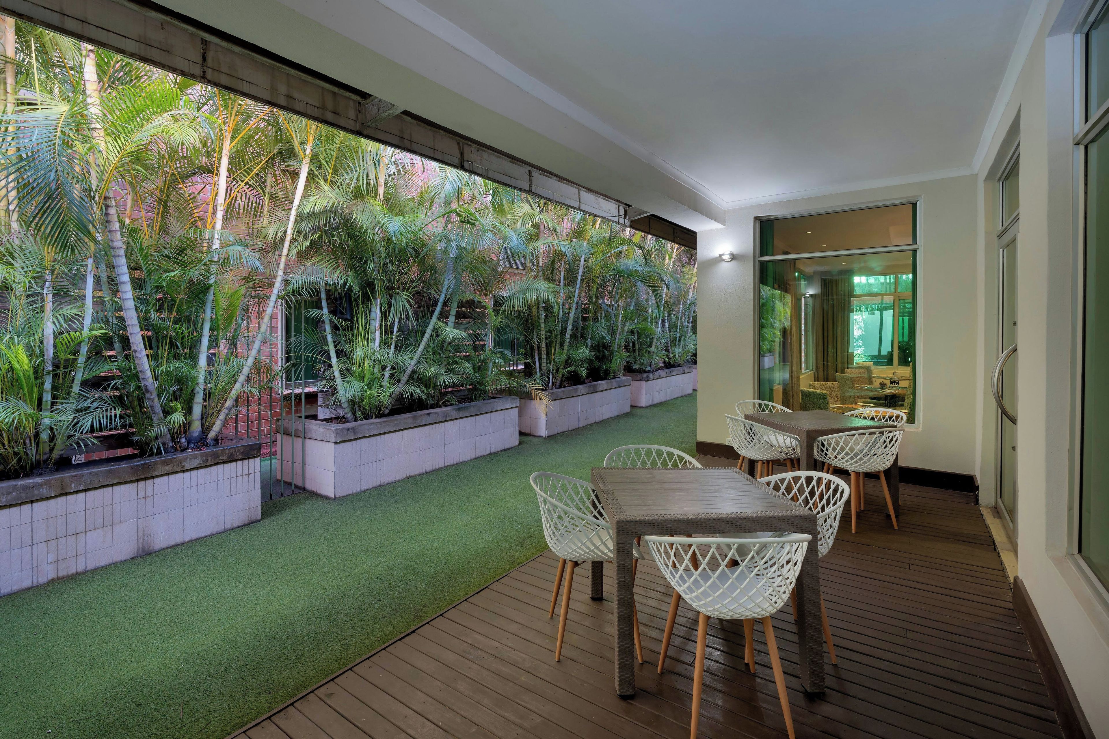 An outdoor patio with a wooden deck, white wicker furniture, and a lush garden with various plants and trees.