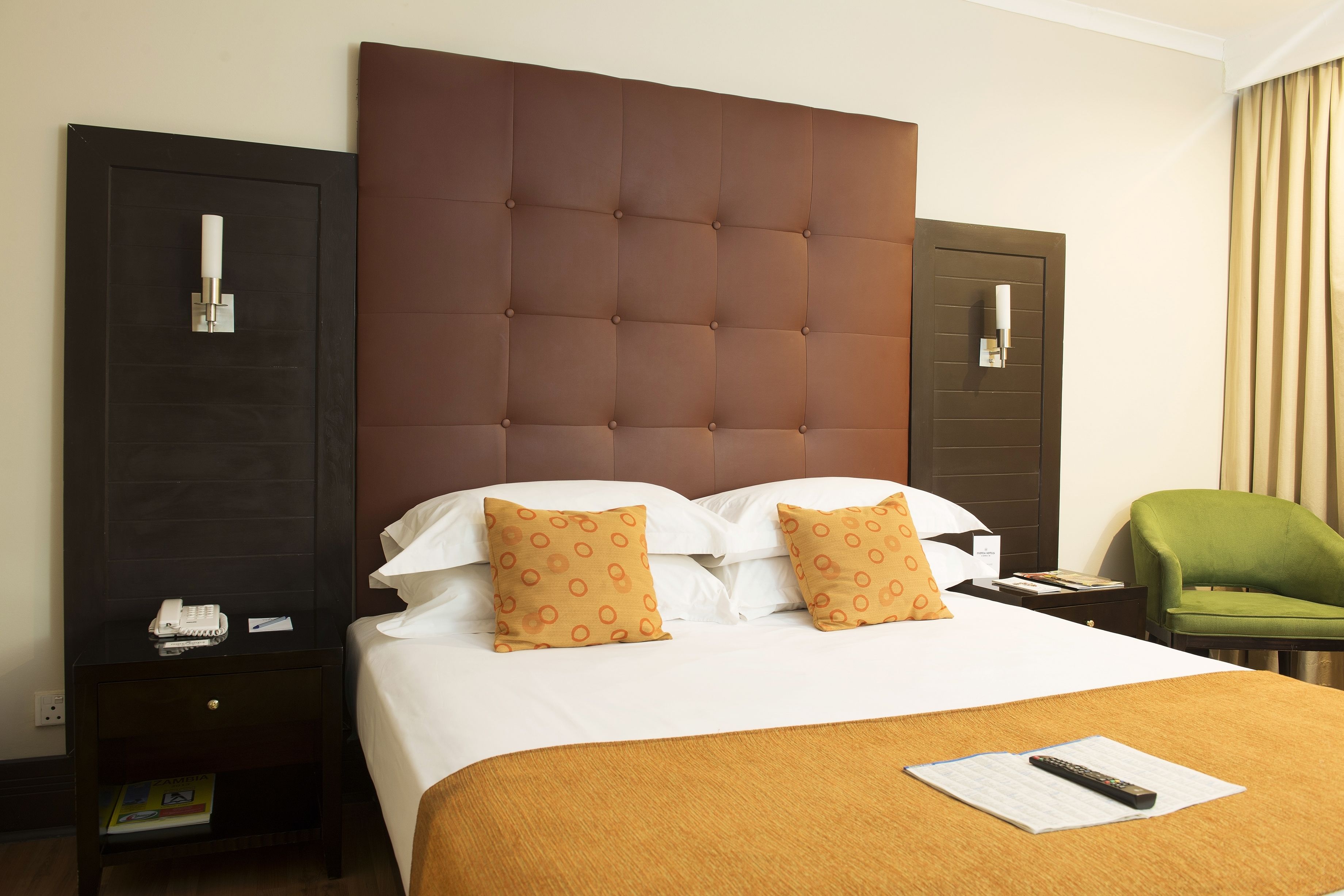 A hotel room with a large bed, two pillows with a polka dot pattern, a green armchair, a nightstand with a telephone, and a remote control on the bed.