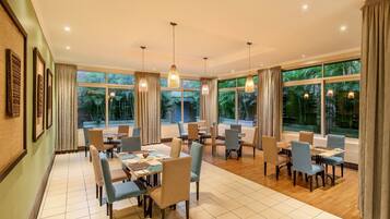 A spacious dining area with multiple tables and chairs, large windows with curtains, and hanging pendant lights.
