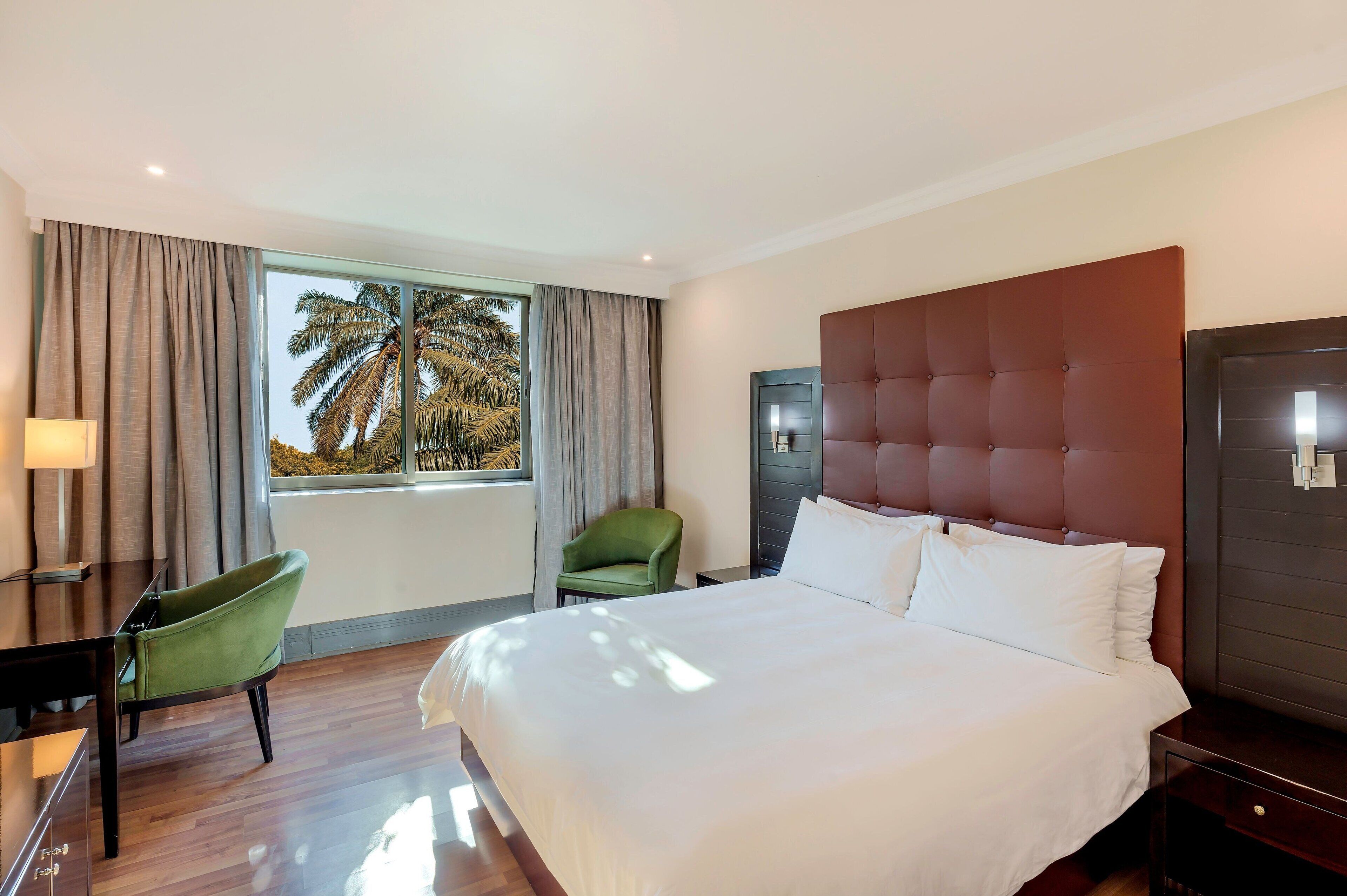 A hotel room with a large bed, a desk, a green chair, and a view of palm trees through the window.