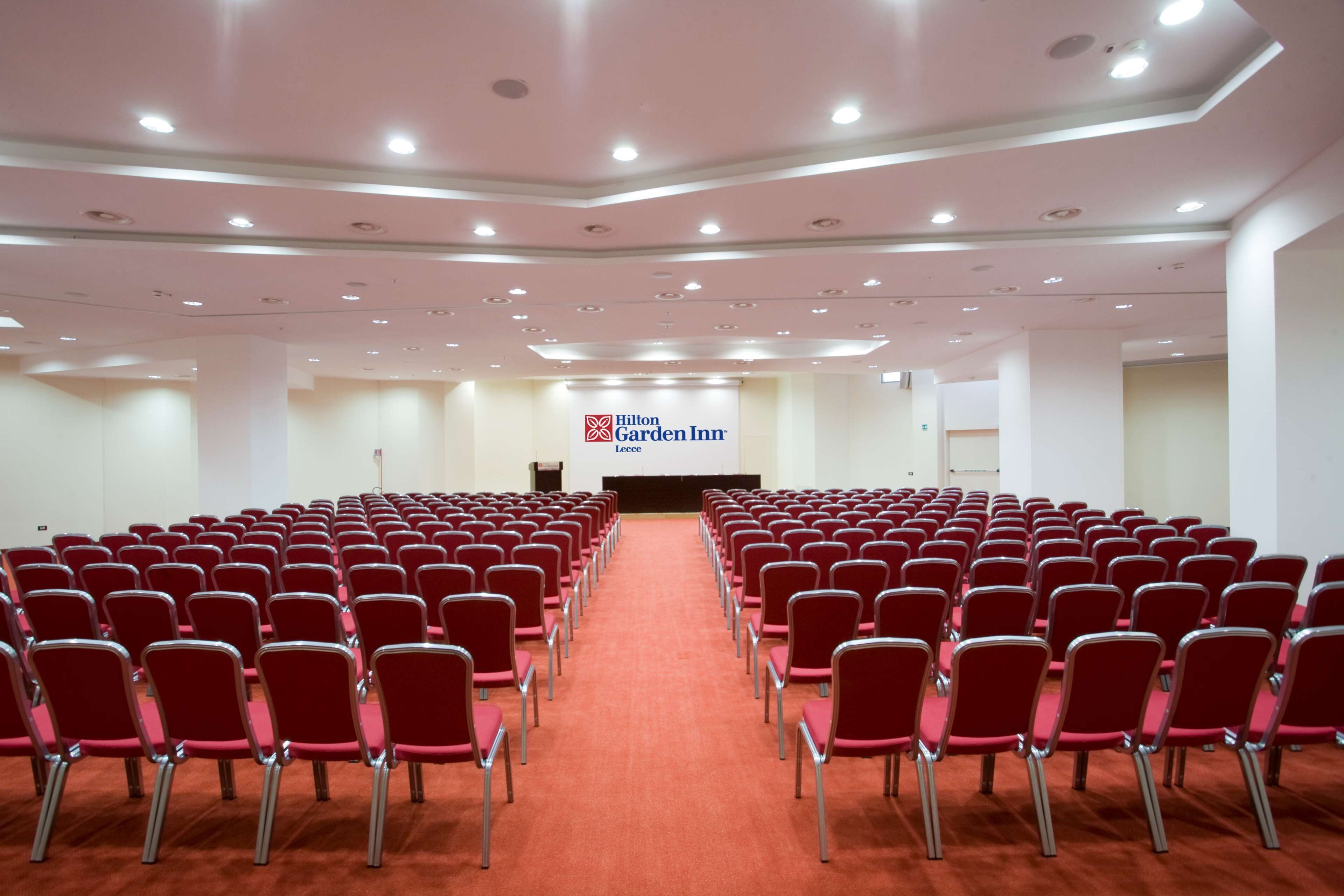 Un salón de conferencias con filas de sillas rojas, un escenario con un atril, y el cartel de Hilton Garden Inn.