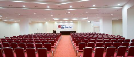 Un salón de conferencias con filas de sillas rojas, un escenario con un atril, y el cartel de Hilton Garden Inn.