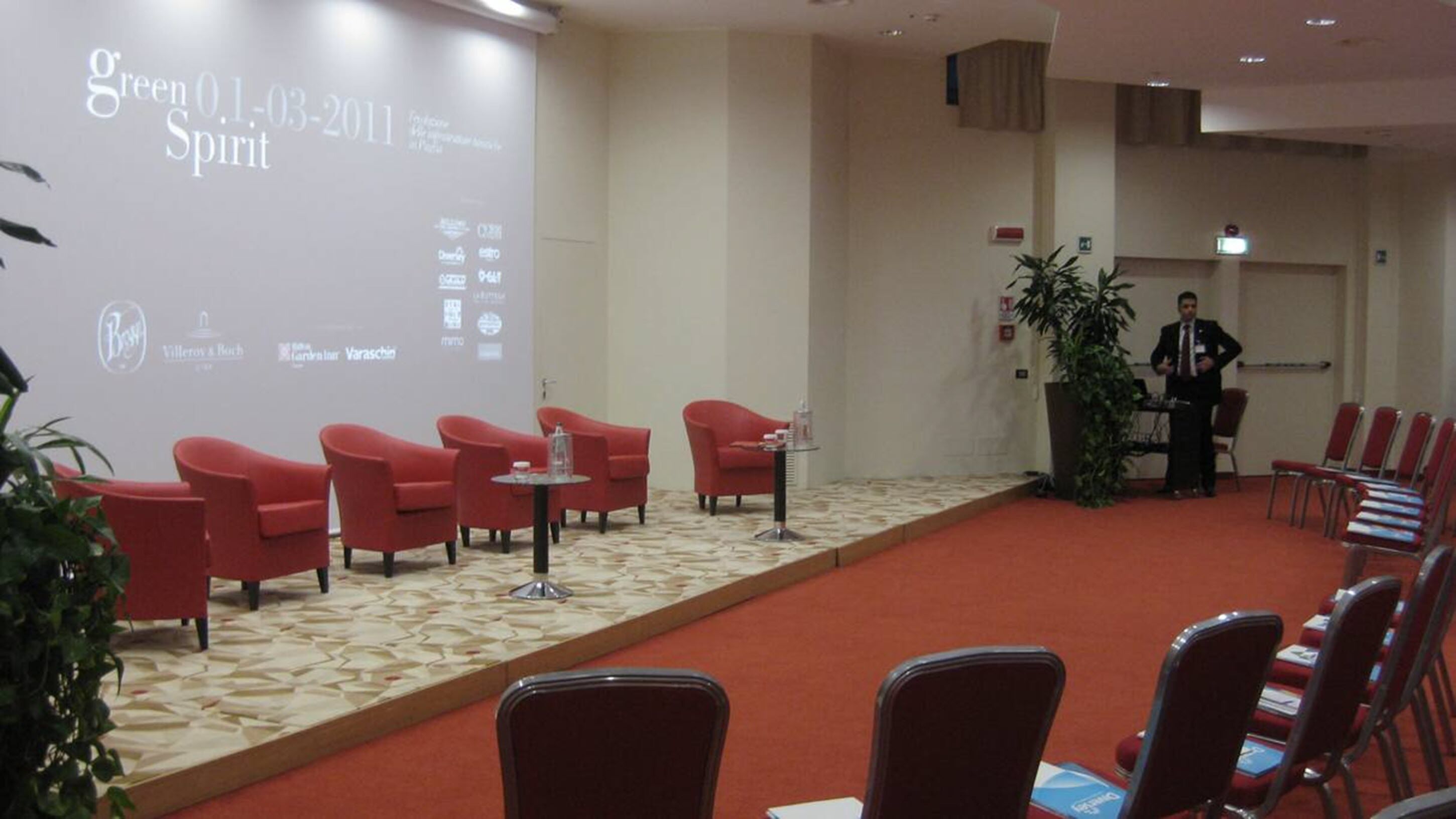 Un salón de conferencias con sillas rojas, piso alfombrado y una pantalla mostrando “green 0.1-03-2011 Spirit”.