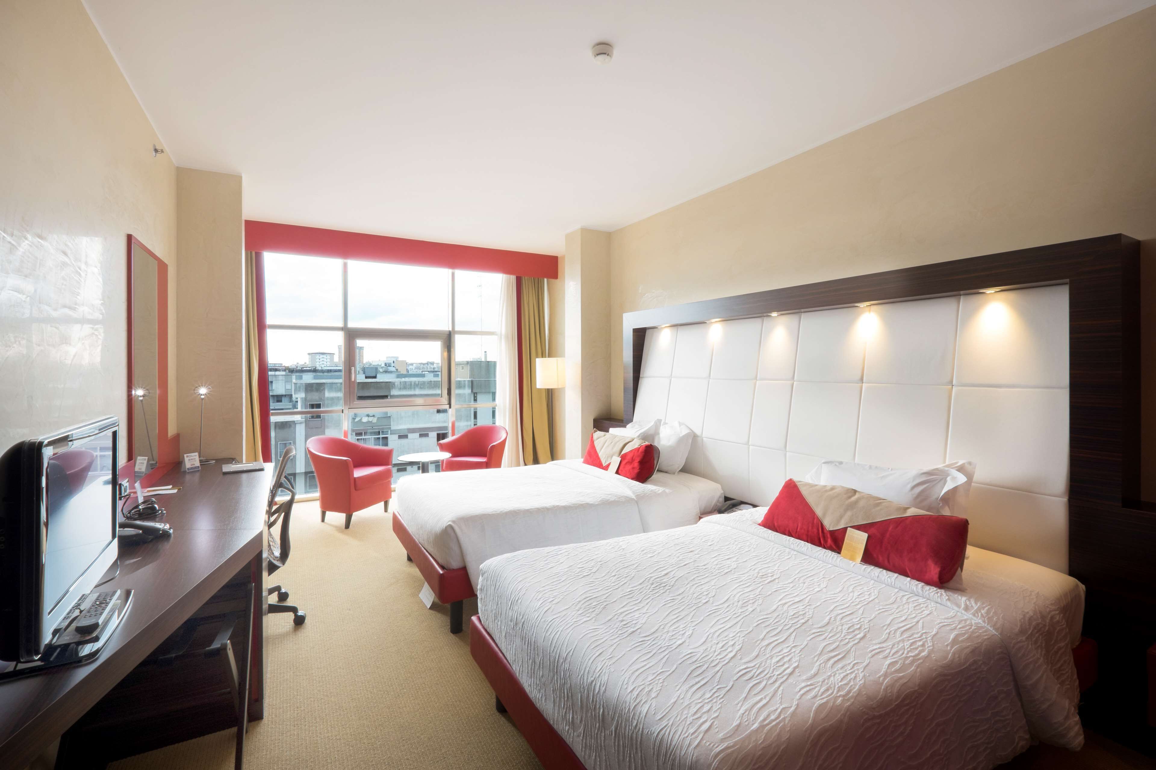 A hotel room with two beds, a desk with a computer, a chair, and a large window offering a city view.