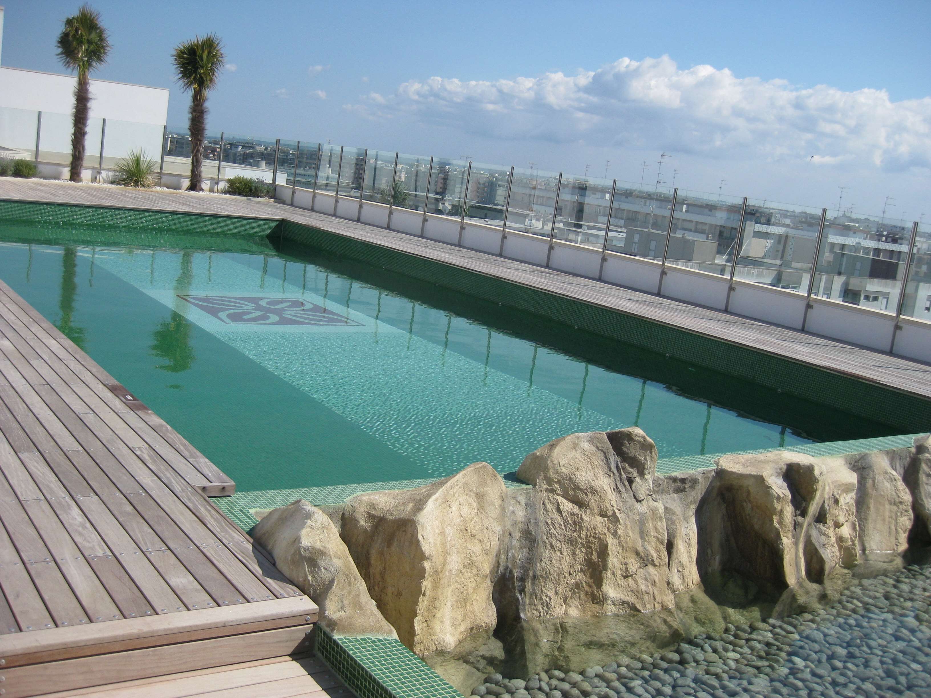 A rooftop swimming pool with clear water, surrounded by a wooden deck and large rocks.
