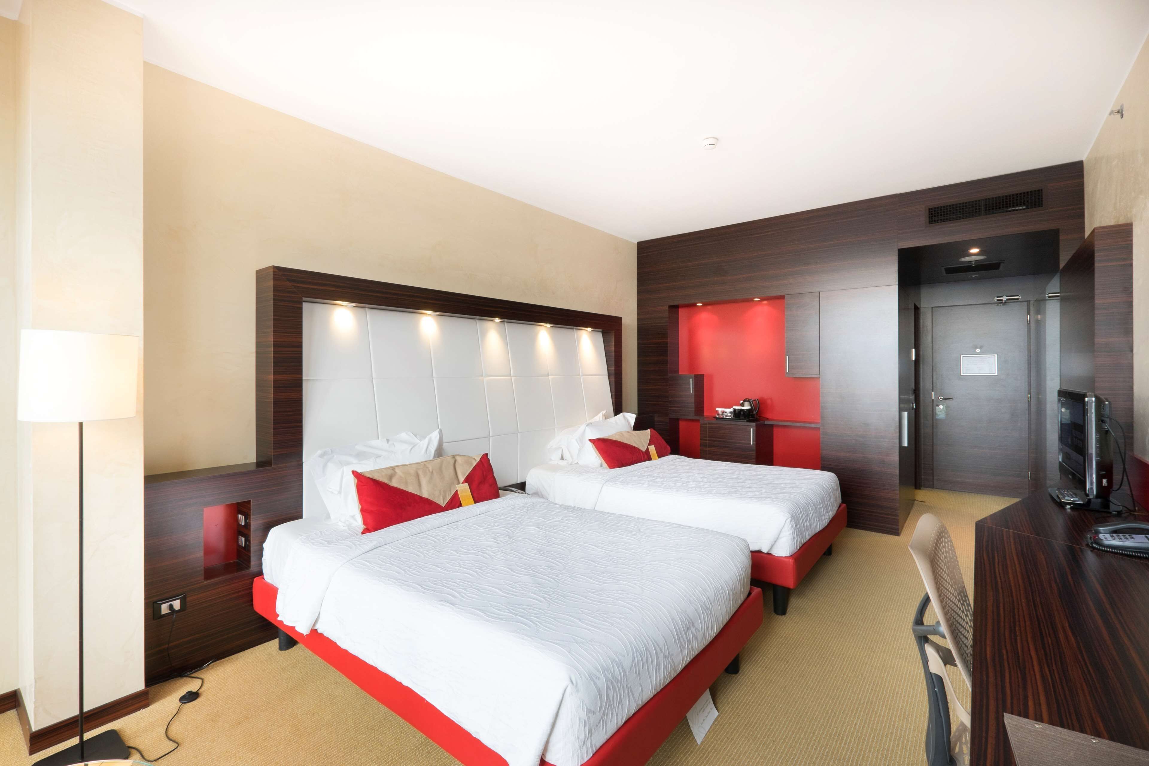 A hotel room with two beds, a desk, a TV, and a red accent wall.