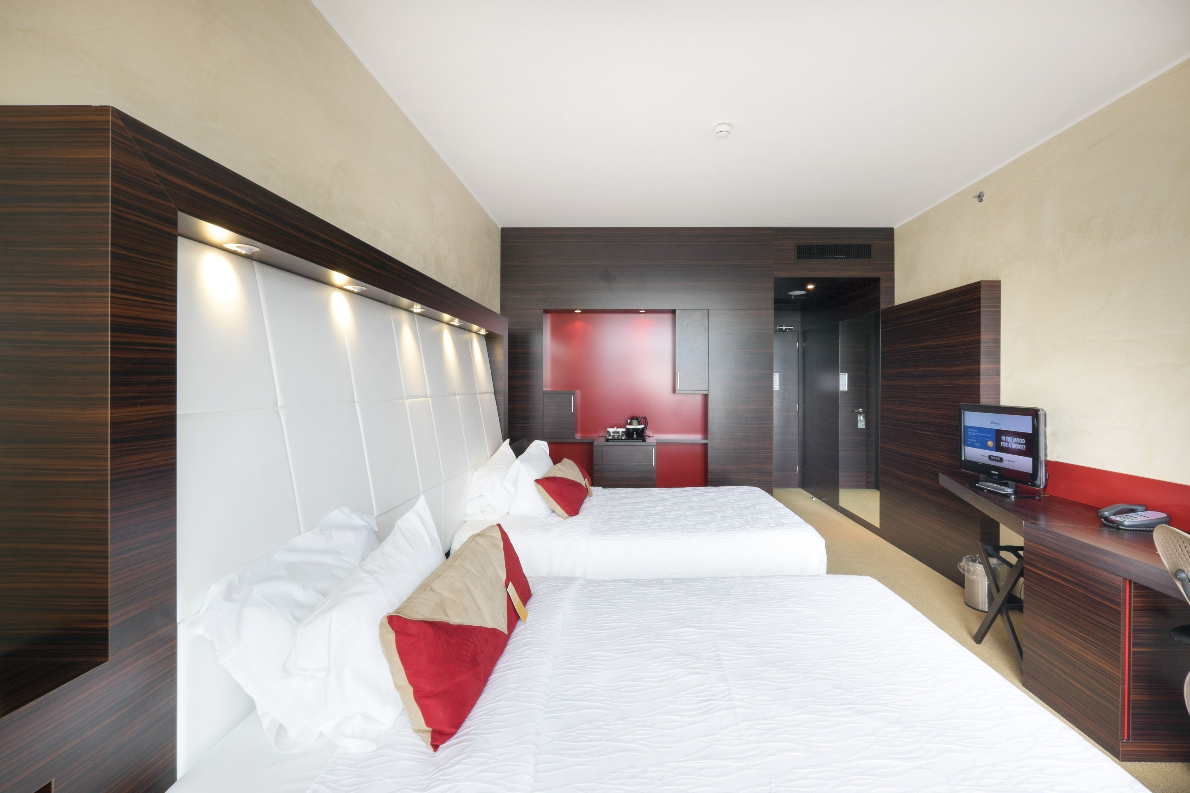 A hotel room with two beds, a desk with a computer, and a red accent wall.
