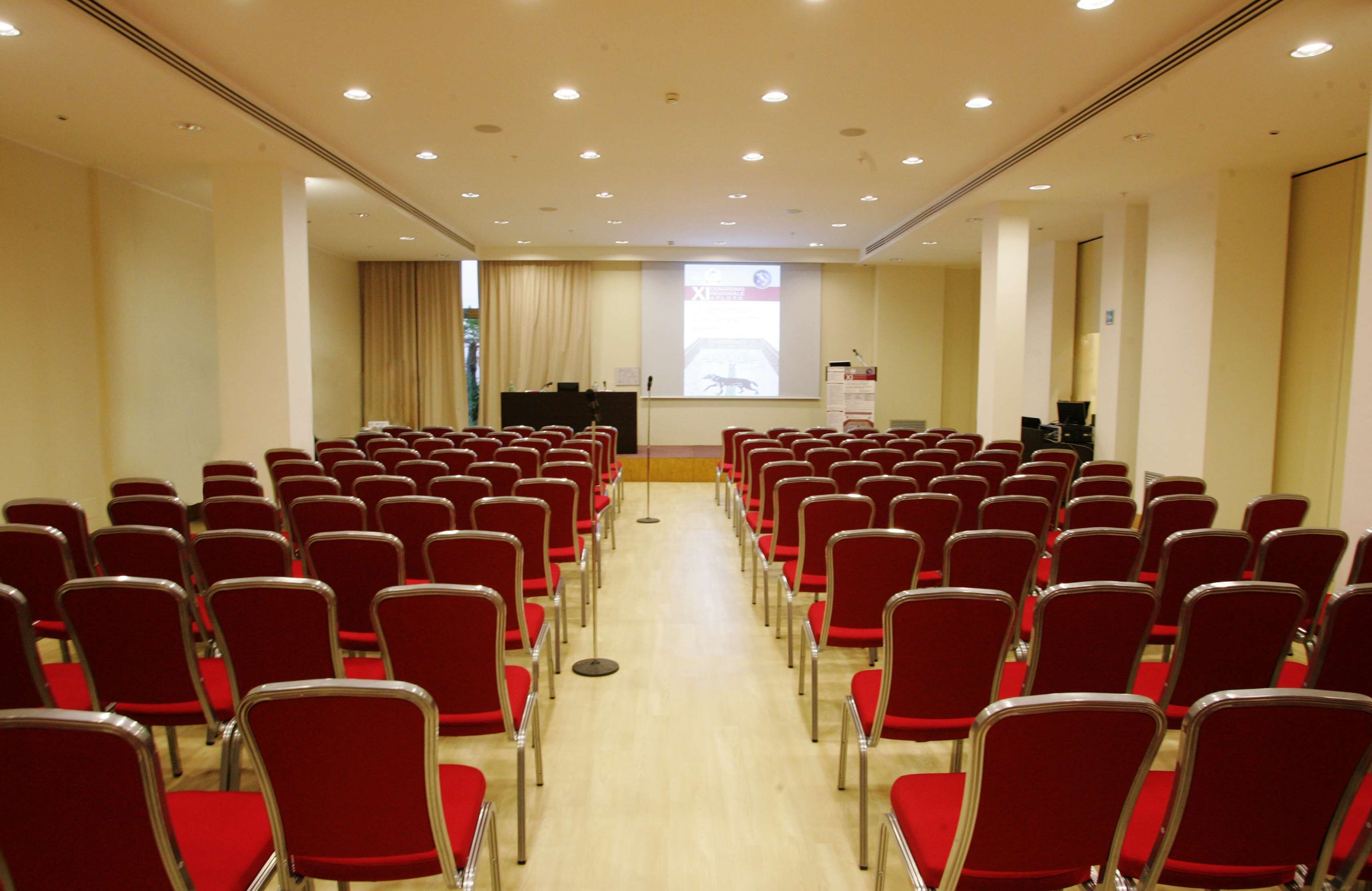A conference room with rows of red chairs, a podium, and a projection screen.