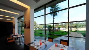 Un restaurante con mesas dispuestas para comer, ventanas amplias que ofrecen vistas a un jardín y una palmera, y una decoración interior moderna.