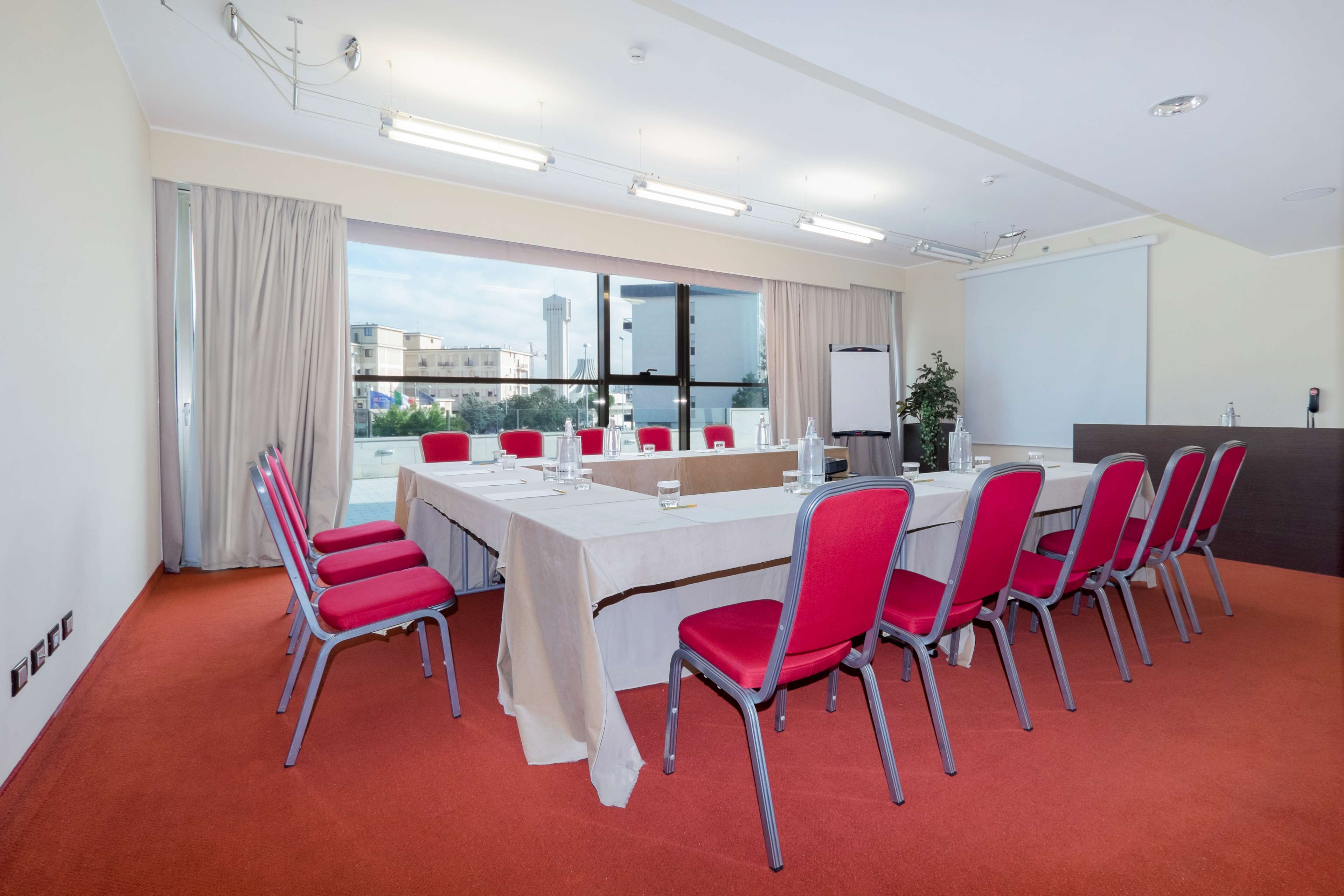 Un salón de conferencias con una larga mesa, sillas rojas, una pizarra y vistas a la ciudad a través de amplias ventanas.