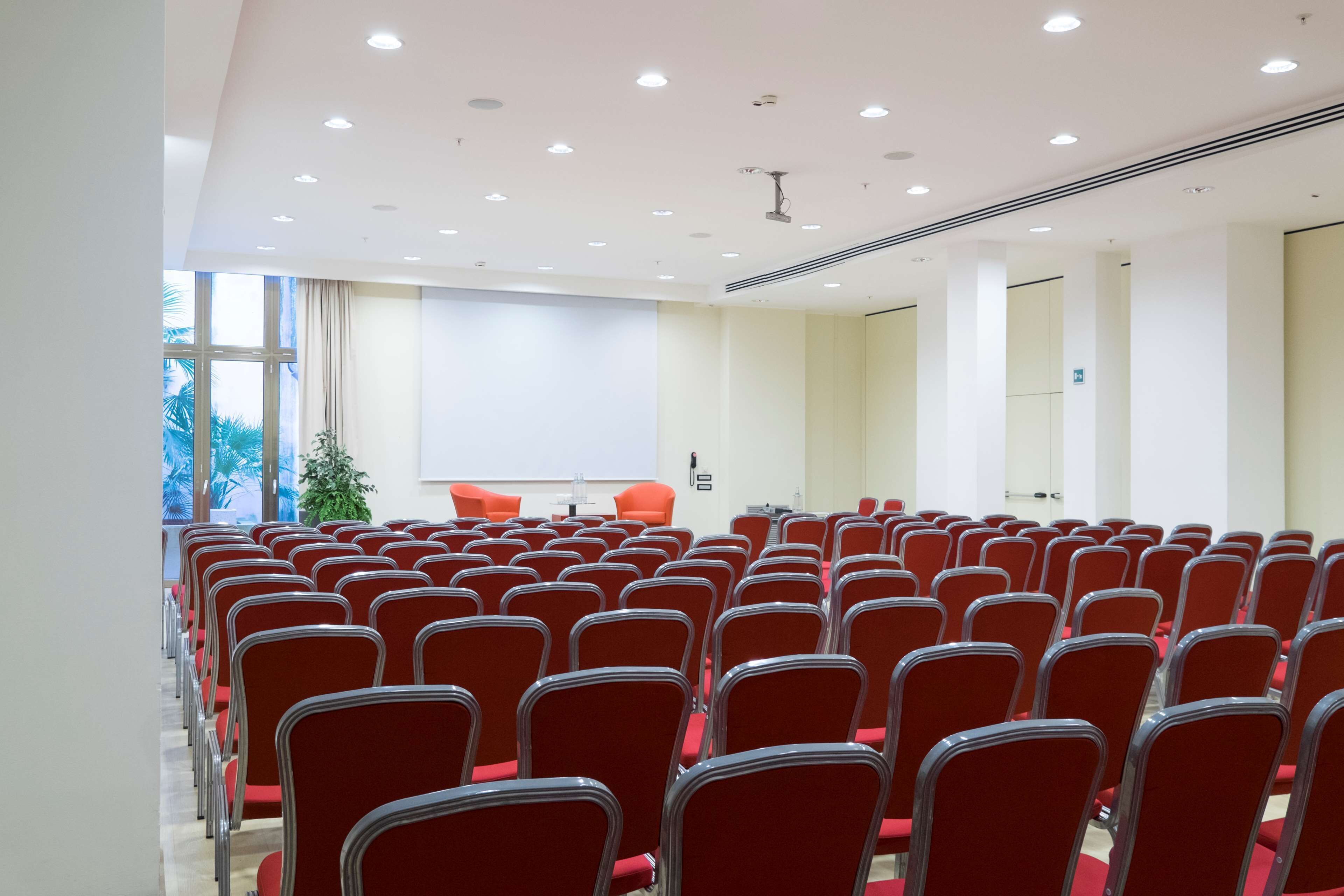 Un salón de conferencias con filas de sillas rojas, una pantalla de proyector y una mesa con sillas naranjas.