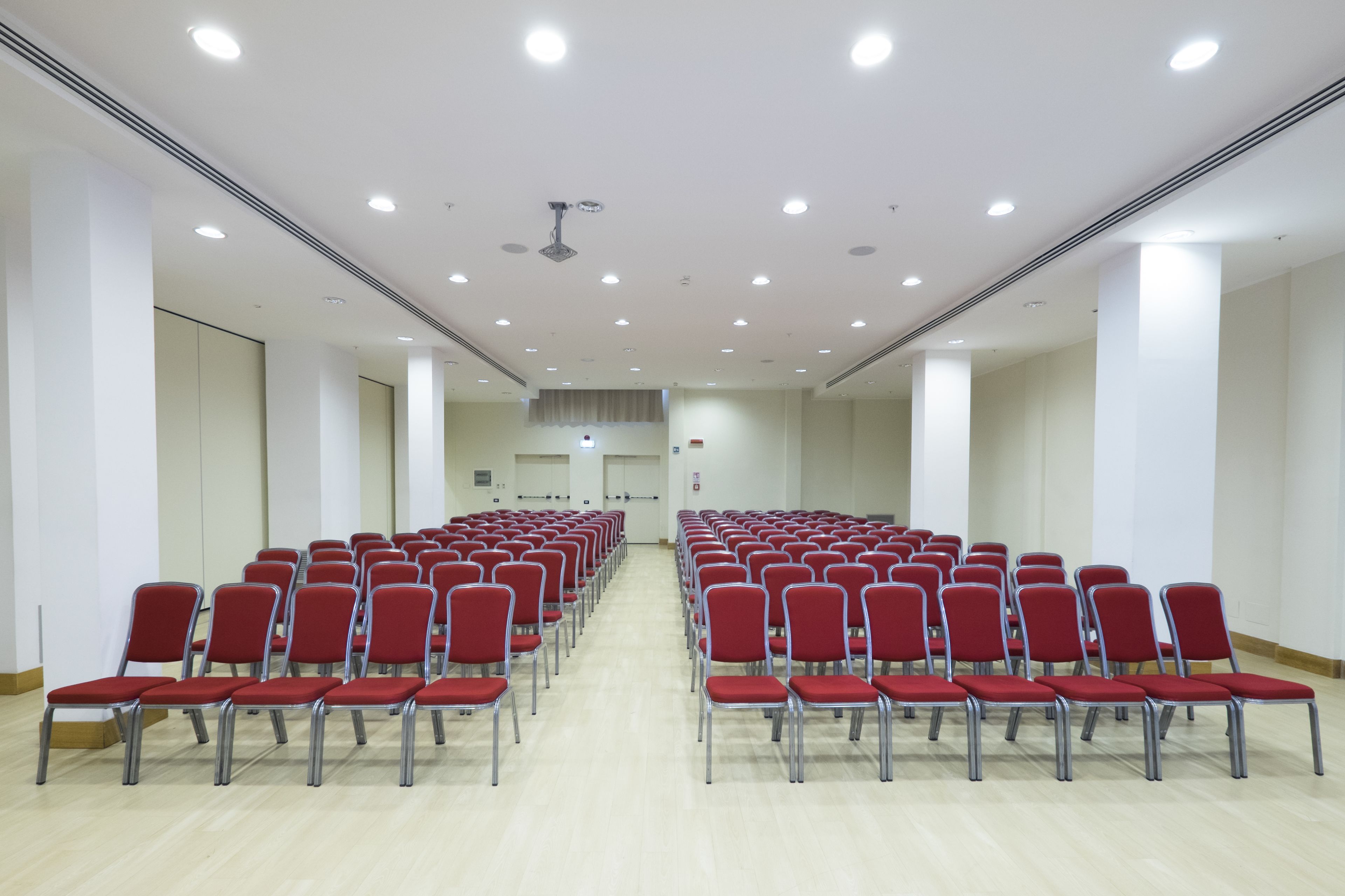 A large, well-lit room with rows of red chairs and a wooden floor, likely a conference or meeting space.