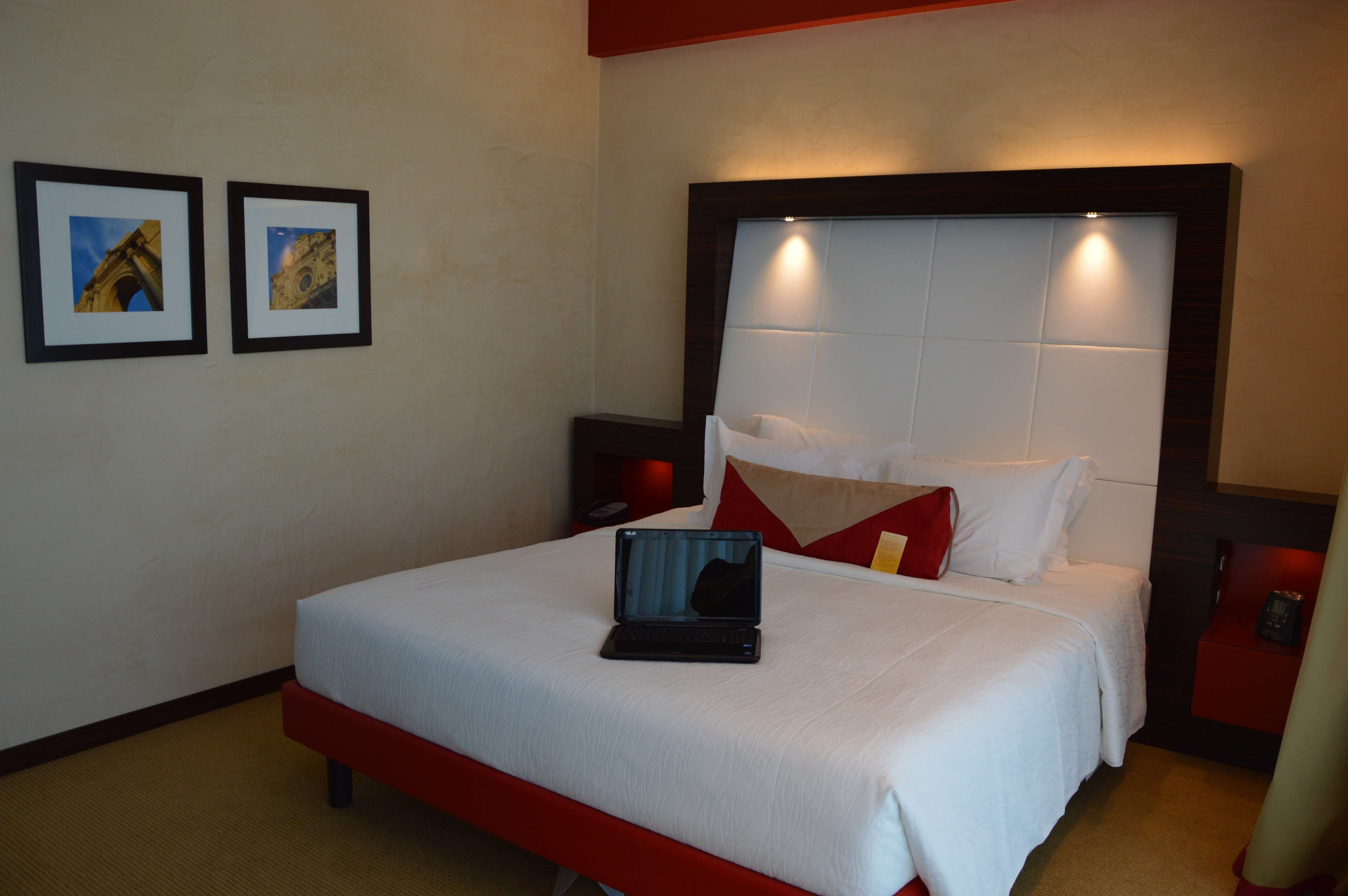 A hotel room with a large bed, a laptop on the bed, two framed pictures on the wall, and a red accent wall.