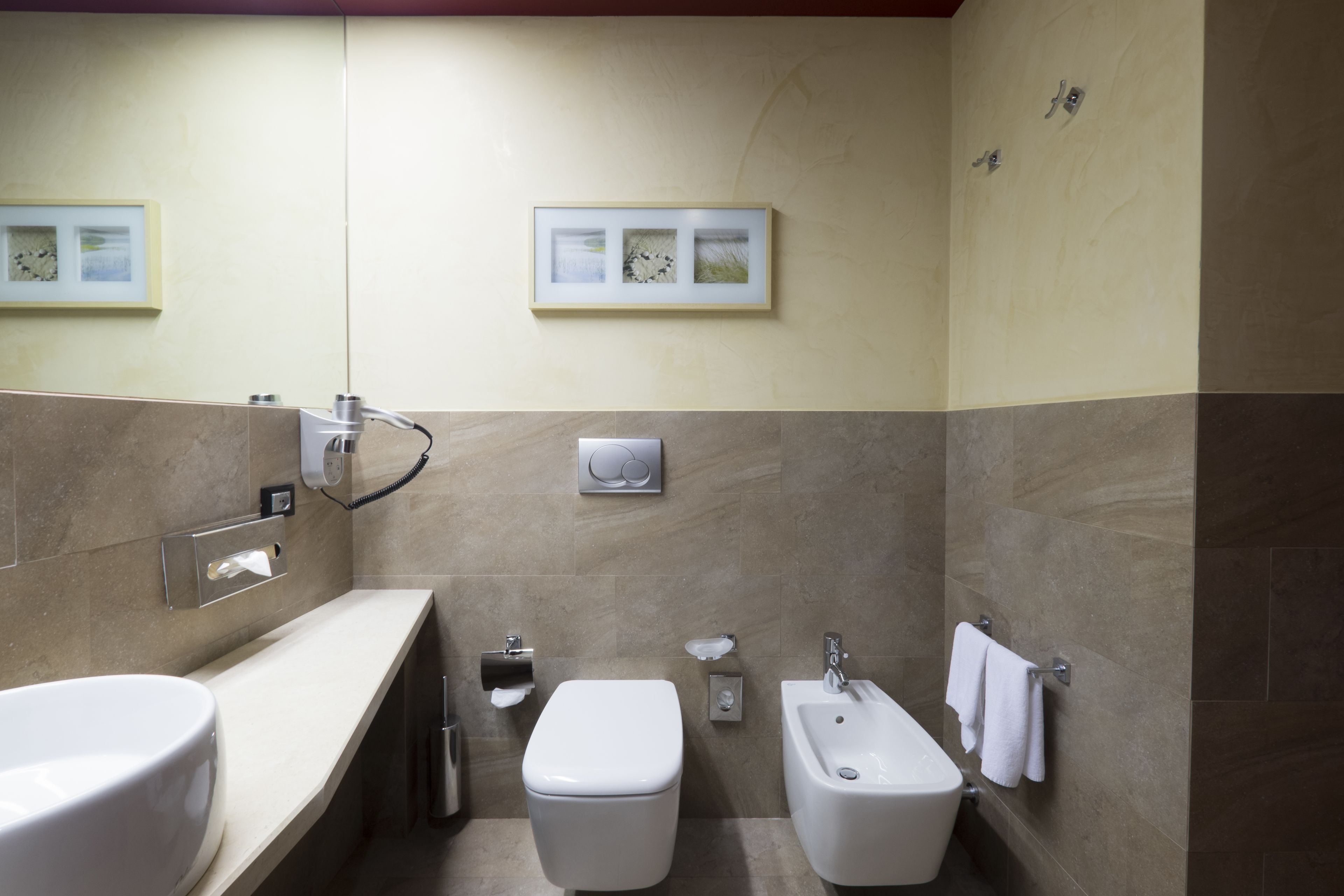 A modern bathroom with a toilet, bidet, and sink. There are framed pictures on the wall and a towel rack.