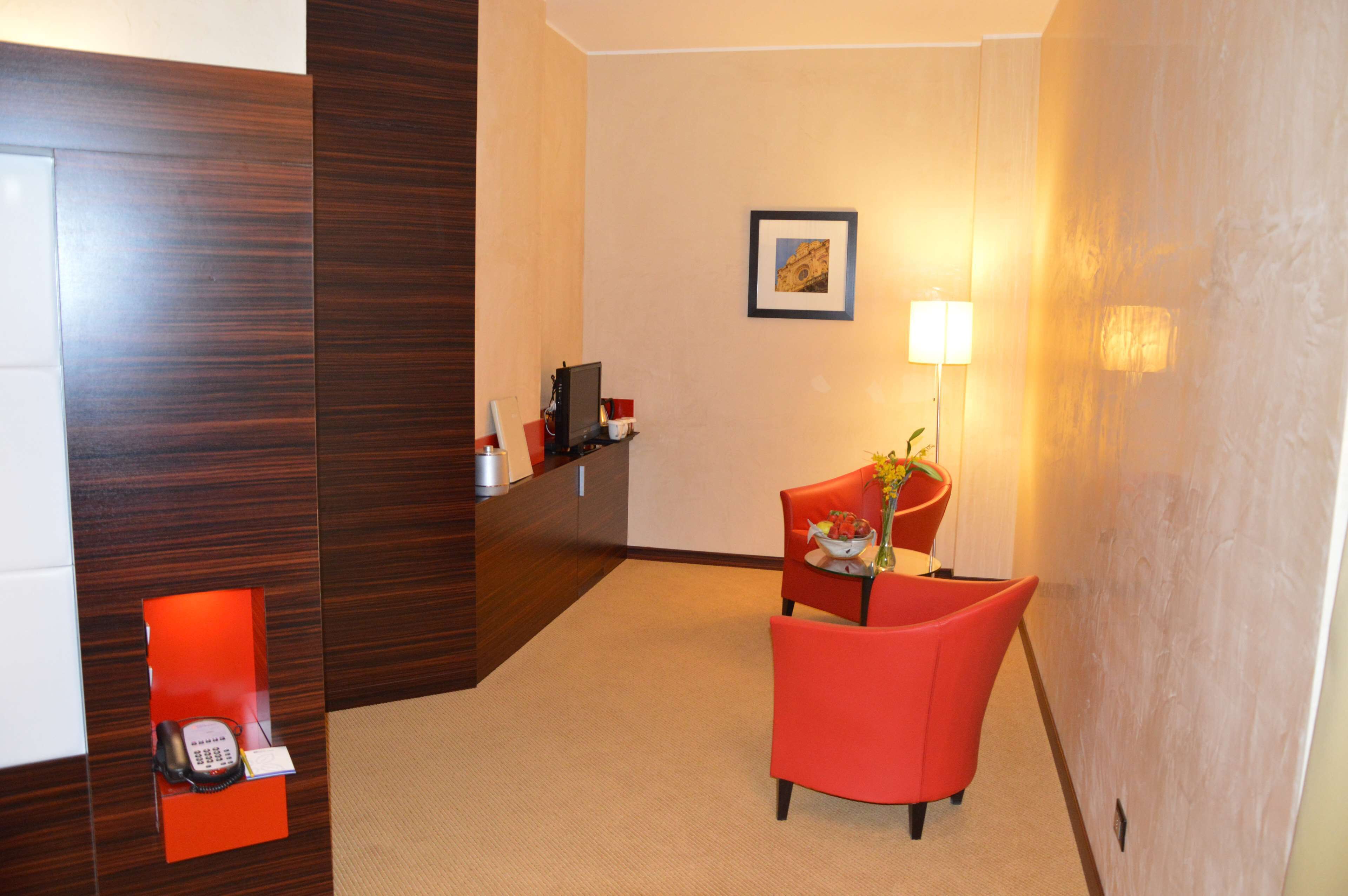 A hotel reception area with a wooden reception desk, a red chair, a small table with a vase and flowers, and a framed picture on the wall.