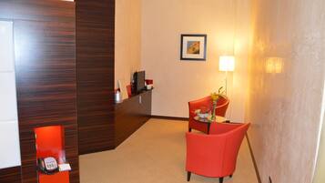Un espace d’accueil d’hôtel avec un comptoir de réception en bois, une chaise rouge, une petite table avec un vase et des fleurs, et un tableau encadré accroché au mur.