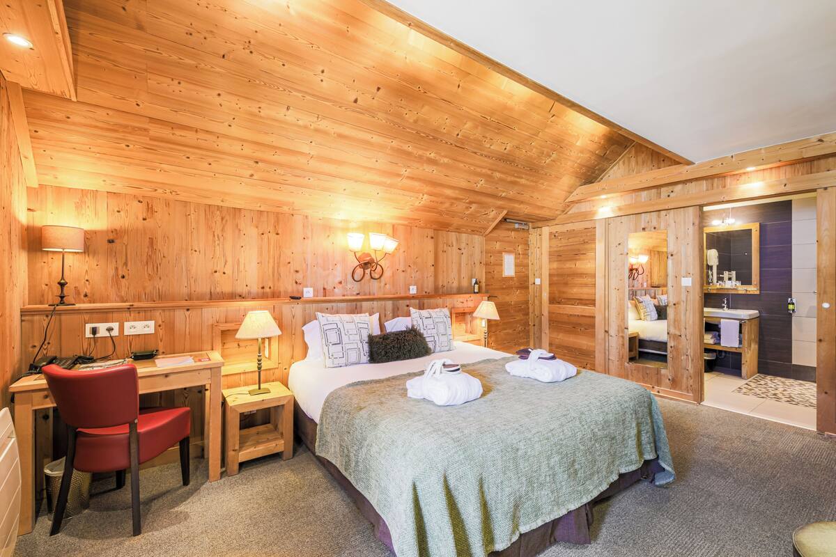 Une pièce aux murs lambrissés, avec un lit, un bureau et une chaise. Une salle adjacente contient un miroir et un lavabo.