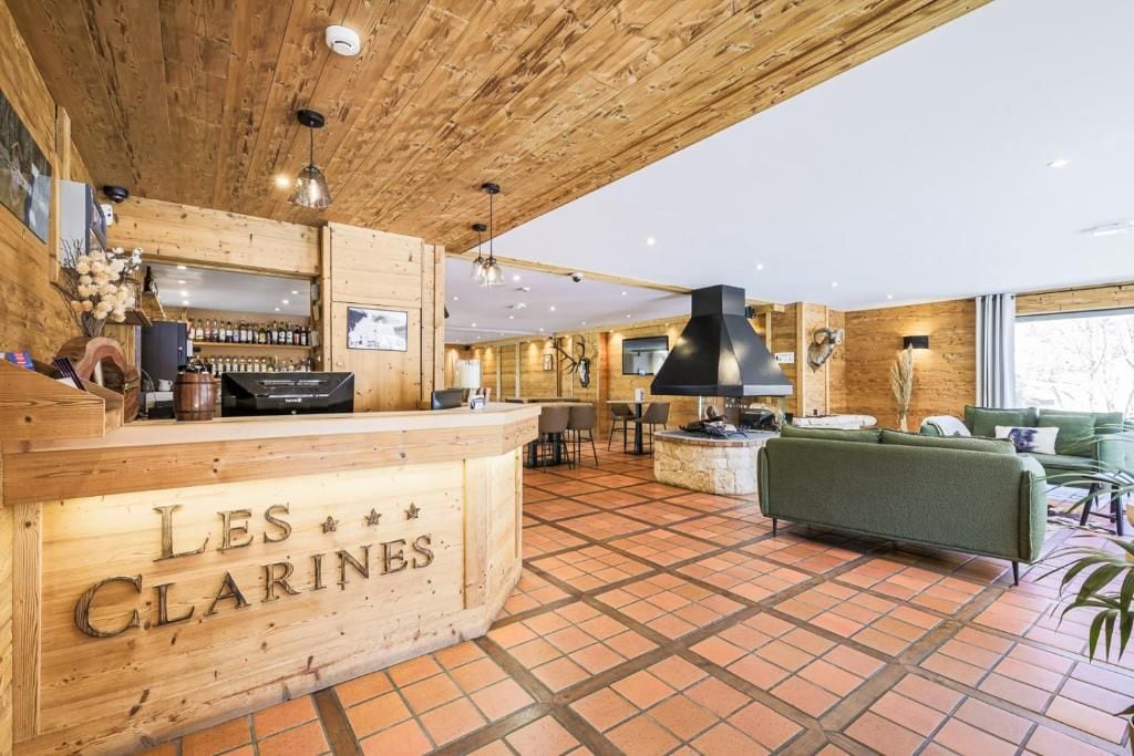 Zona de recepción con mostrador de madera que indica 'LES CLARINES', bar con botellas, chimenea y sofá verde.