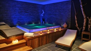 Een wellnessruimte met een stenen muur, een jacuzzi, houten ligbedden en kaarsen.