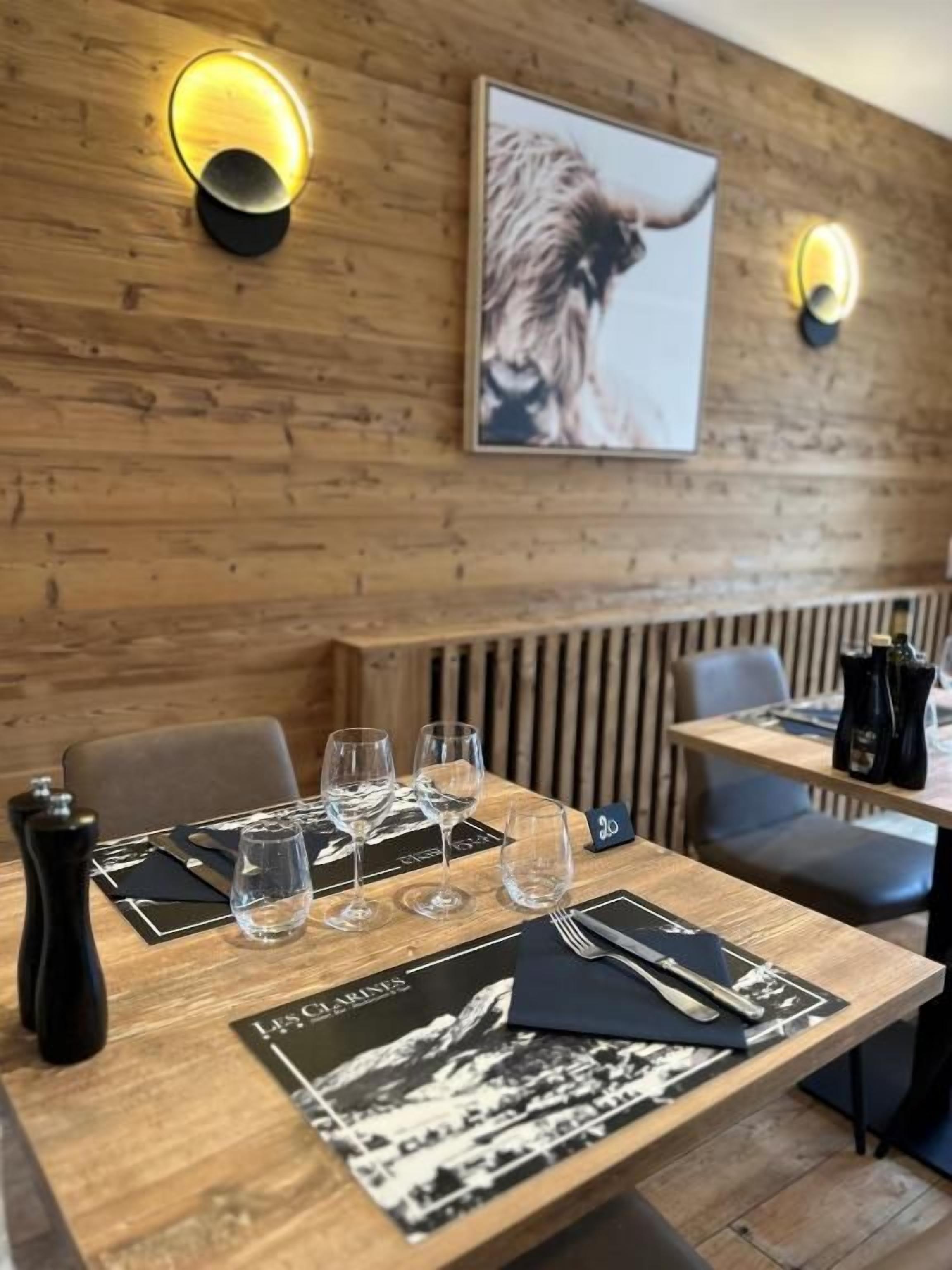 Een restaurantinterieur met houten wanden, twee wandlampen, een gedekte tafel met wijnglazen, een menukaart en een ingelijst schilderij aan de muur.