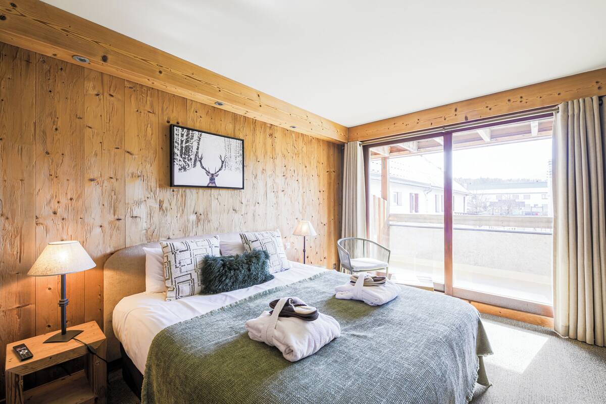 Une chambre d’hôtel avec un mur en bois, un lit recouvert d’une couverture verte, une chaise et une fenêtre avec des rideaux.