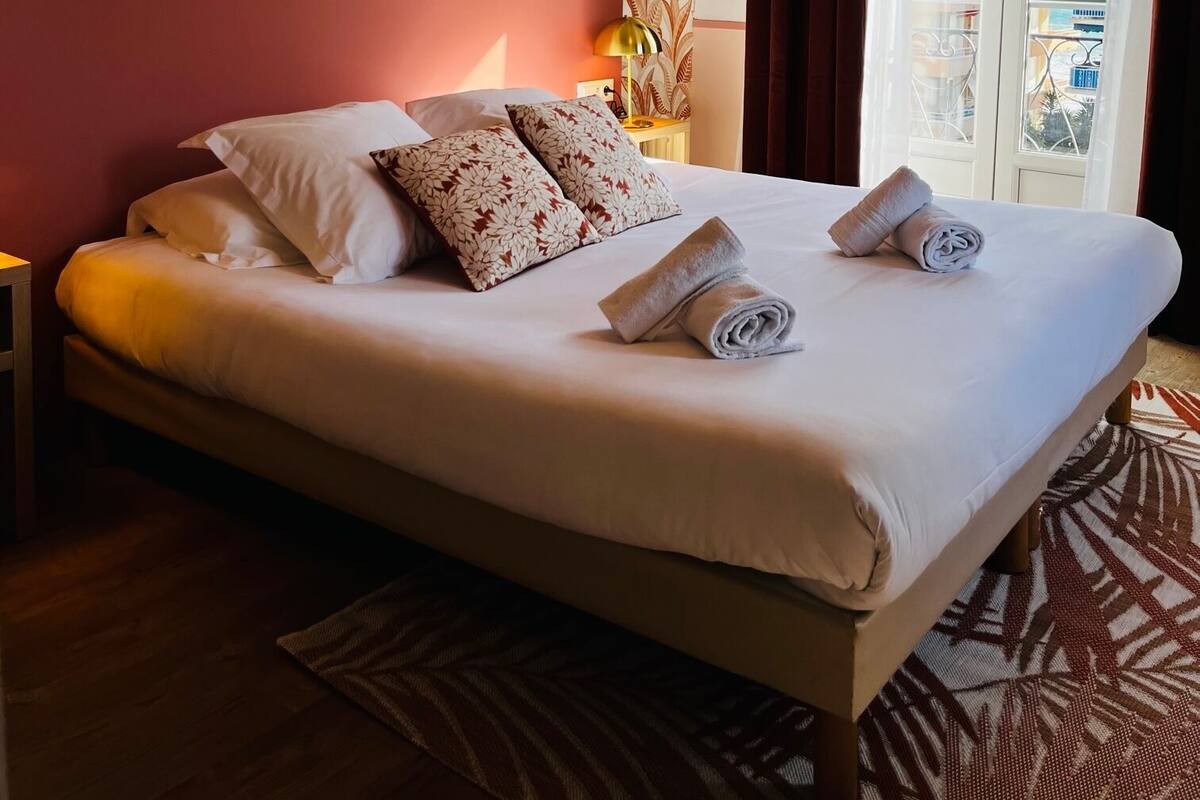 A hotel room with a large bed, floral wallpaper, a view of buildings through the window, and a nightstand with a lamp.