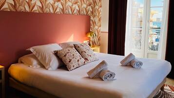 A hotel room with a large bed, floral wallpaper, a view of buildings through the window, and a nightstand with a lamp.